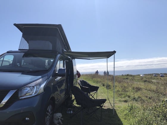 Campervan with awning and camping chairs out on grass overlooking sea under clear blue sky in Cornwall