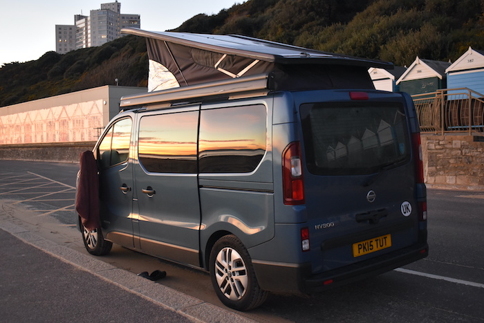 blue campervan with reflection of sunset and beach huts in windows